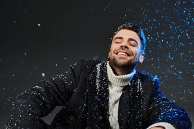 Yakışıklı bir genç adam, kış atmosferinde yumuşak kar taneleriyle çevrili, mutlu bir şekilde oturur..