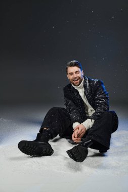Bu şık kış kıyafetleri içindeki genç adam karda neşeyle oturur, mutluluk ve cazibe saçar..