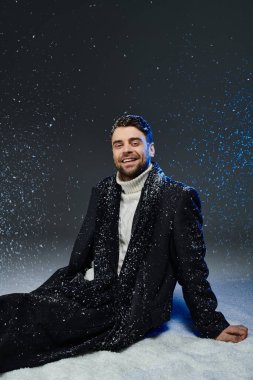 Çekici genç bir adam yerde dinleniyor, etrafında kar taneleri uçuşurken neşeyle gülümsüyor..
