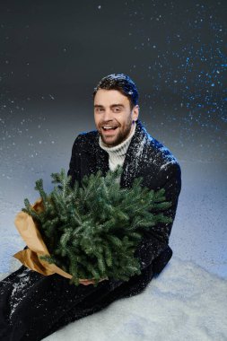 Neşeli genç bir adam, düşen kar taneleriyle çevrili bir Noel ağacını kucaklıyor..