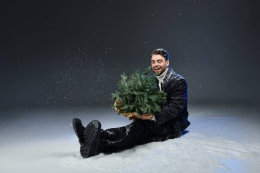 Genç adam karla çevrili küçük bir Noel ağacını kucaklarken ışıl ışıl gülümsüyor..