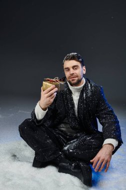 A young man smiles while sitting in the snow, admiring a festive gift wrapped in gold. clipart