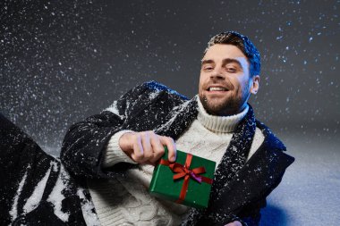 Neşeli genç bir adam güzel paketlenmiş bir hediyeyi sunarken karlı bir dekor içinde uzanır..