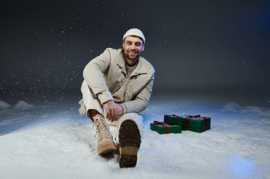 Genç adam neşeli bir şekilde karın üzerinde otururken etrafı hediyelerle çevriliyken gülümsüyor..