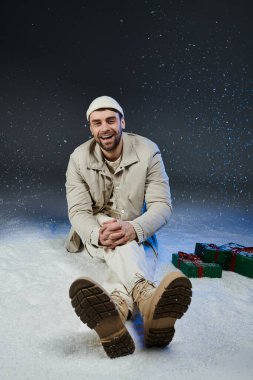 Neşeli genç bir adam yumuşak karda oturur, etrafını renkli hediyeler sarar, mutluluk saçar..
