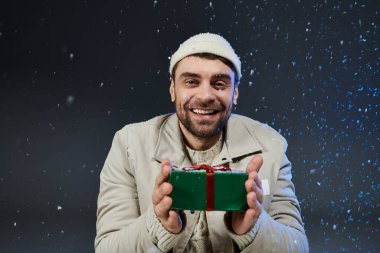 Kış kıyafetleri içinde neşeli bir genç adam kar yağarken bir hediye tutar..