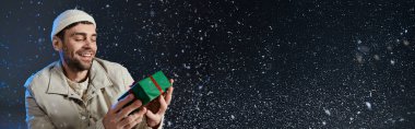Genç bir adam renkli bir hediyeyi açarken ışıl ışıl gülümsüyor, karlı atmosferin tadını çıkarıyor..