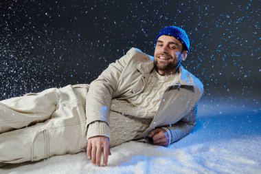 A cheerful young man relaxes in the snow, smiling joyfully as snowflakes fall around him. clipart