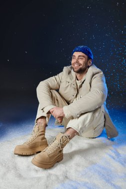 The young man smiles joyfully while sitting in the snow, dressed in cozy winter attire. clipart