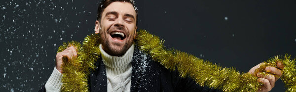 A cheerful young man joyfully wraps himself in festive decorations while snow falls around.