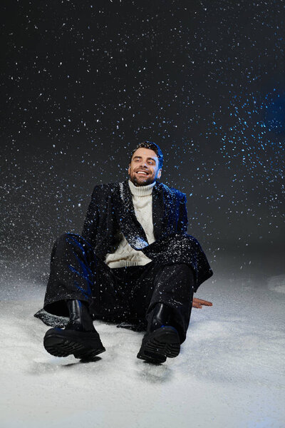 A charming young man smiles joyfully while sitting in snow, surrounded by swirling flakes.