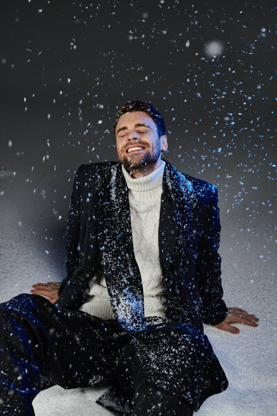 The young man sits in a flurry of snow, smiling joyfully in his stylish winter outfit.