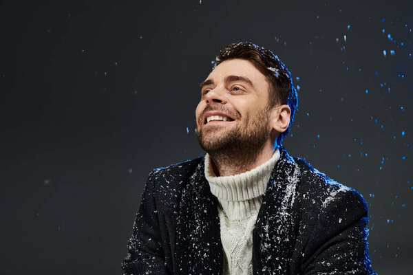 A cheerful young man smiles joyfully as snowflakes gently fall around him in a cozy setting.