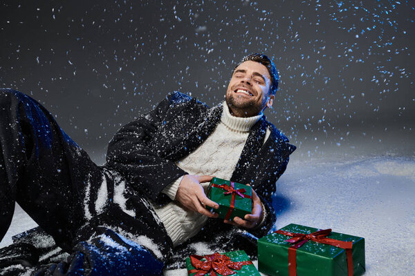 Веселый молодой человек сидит на заснеженной земле в окружении подарков, наслаждаясь моментом.