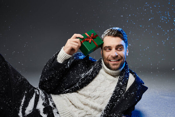 Красивый молодой человек улыбается, держа в руках завернутый подарок, окруженный падающими снежинками.
