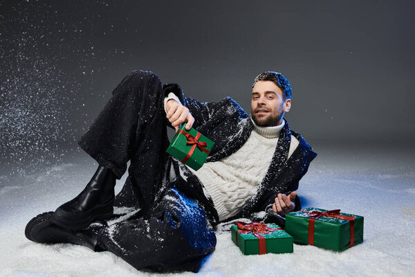Красивый молодой человек расслабляется на снежном фоне, игриво держа завернутые праздничные подарки.