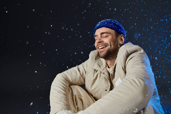 A joyful young man embraces the snowflakes as he relaxes in winter attire, radiating happiness.