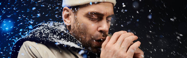 A handsome young man blows warmth into his hands, surrounded by swirling snowflakes.