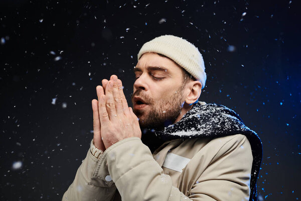 A stylish young man is warming his hands together against a backdrop of falling snowflakes.