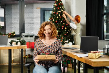 Arkaplanda Noel ağacını süsleyen iş arkadaşıyla birlikte hediyeyle gelen genç bir kadın..