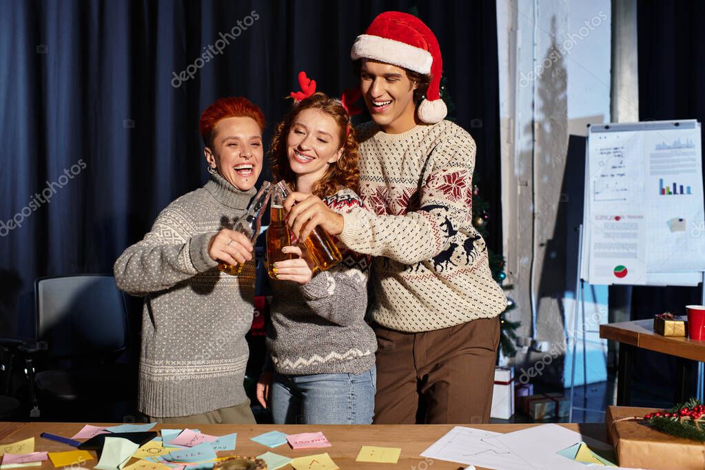Three cheerful coworkers in Christmas sweaters raise drinks together at a holiday office party.