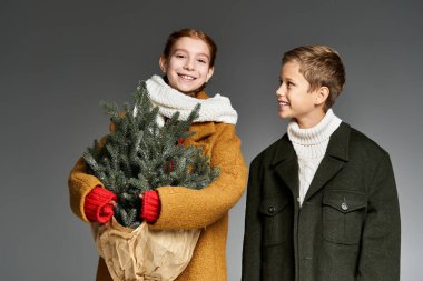 Two cheerful kids dressed warmly smile while carrying a tiny Christmas tree wrapped in brown paper. clipart