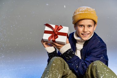 A cheerful boy in winter clothes smiles while presenting a wrapped gift. clipart