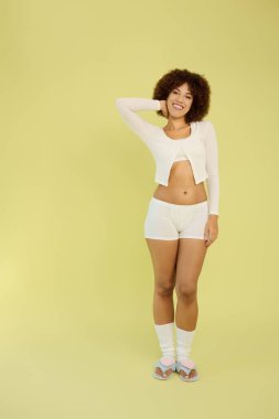 Kıvırcık saçlı genç bir kadın, parlak sarı bir arka planda iç çamaşırlarıyla güvenle duruyor..