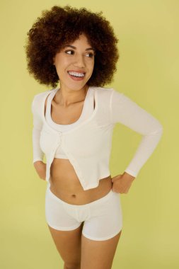 Kıvırcık saçlı genç bir kadın, canlı sarı bir arka planda iç çamaşırıyla dururken mutluluk saçar..