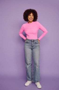 Pembe bluzlu, kıvırcık saçlı ve açık renk kotlu genç bir kadın canlı mor bir arka plana karşı neşe veriyor..