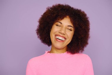 A young woman in pink casual clothing radiates joy with her bright smile, set against a vibrant purple background. clipart