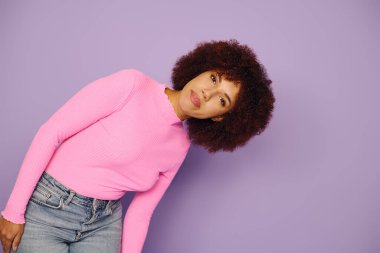 Kıvırcık saçlı genç bir Afro-Amerikalı kadın mor bir arka planda günlük pembe kıyafetler içinde poz veriyor..