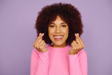 Pembe elbiseli genç bir Afrikalı Amerikalı kadın pırıl pırıl gülümsüyor ve canlı mor bir fon karşısında parmaklarıyla kalpleri gösteriyor..