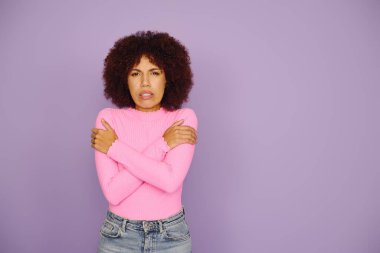 A young African American woman in pink casually expresses her feelings while standing and shivering on a purple background. clipart