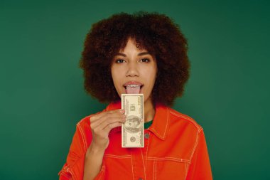 A young African American woman with curly hair shows excitement while holding a dollar bill against green. clipart