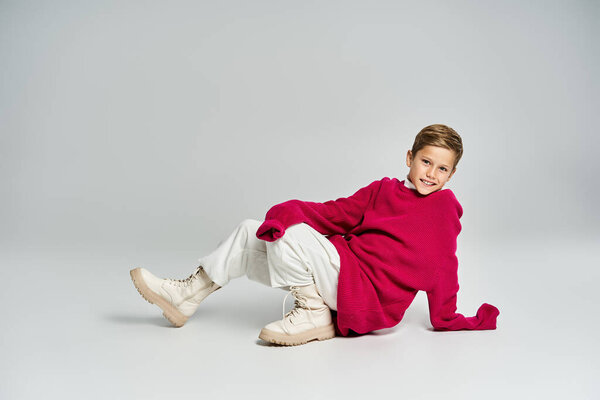 Веселый мальчик в уютной зимней одежде игриво сидит на мягком фоне, излучая тепло.