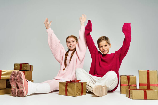 Двое веселых детей, одетых в теплый зимний наряд, наслаждаются своим временем в окружении праздничных подарков.