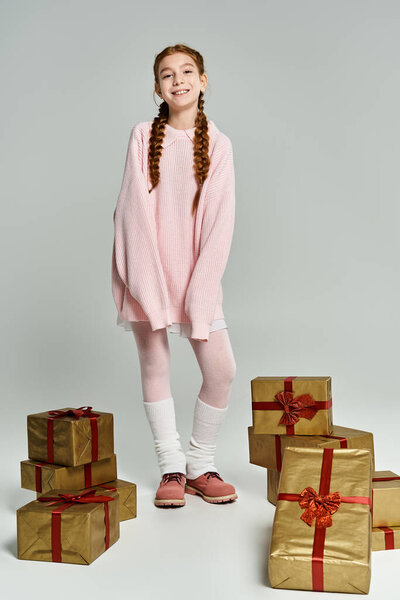 Радостная девушка в уютном розовом наряде стоит среди золотых завернутых подарков, излучающих тепло и счастье.
