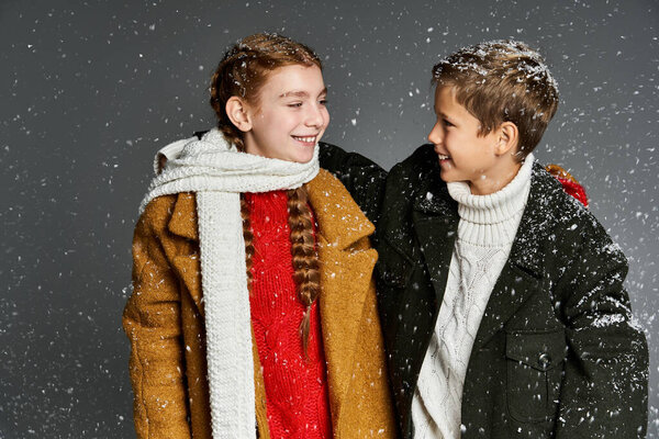 Two smiling children enjoy a winter day dressed in warm, colorful clothing as snowflakes fall.