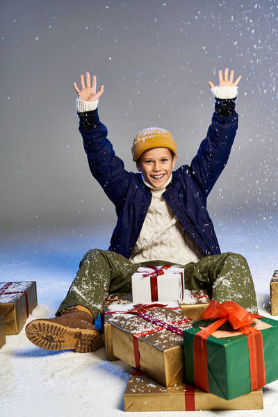 Улыбающийся мальчик в зимнем снаряжении сидит на земле, окруженный красочными подарками в снегу.