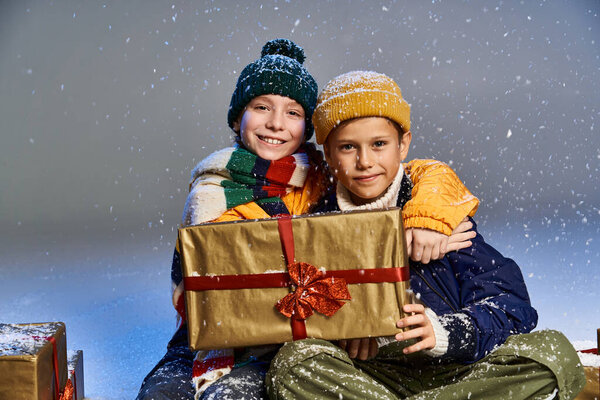 В уютной зимней обстановке двое счастливых детей обнимаются в окружении праздничных подарков.