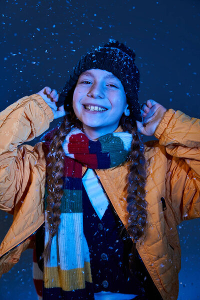 A cheerful girl plays in the snow, wearing a warm jacket and a vibrant scarf while smiling.