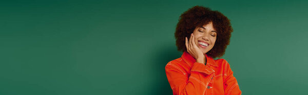 Веселая молодая женщина с вьющимися волосами демонстрирует свои эмоции в повседневной одежде, стоя у зеленой стены.