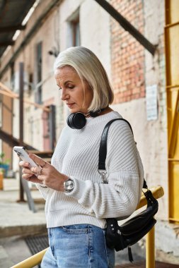 A stylish older woman with gray hair checks her smartphone while leaning against a railing. clipart