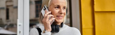 A mature woman with beautiful gray hair smiles as she chats on her phone outside a vibrant building. clipart