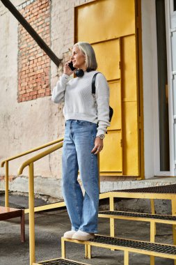 A gray haired woman stands on steps, chatting animatedly on the phone in casual attire. clipart