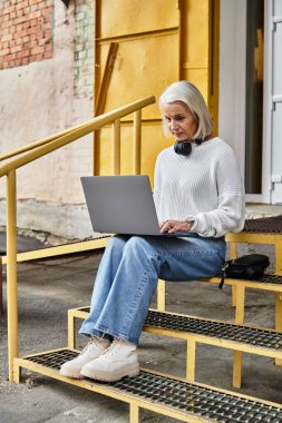 Gri saçlı, güzel, olgun bir kadın laptopuyla dışarıda çalışıyor..