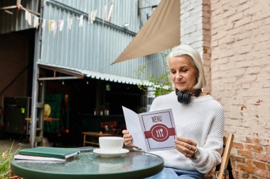 A gray haired woman sits outside, reading a menu with a warm drink nearby. clipart