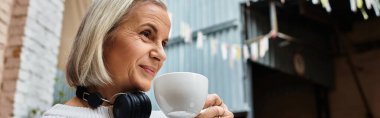 A gray haired woman enjoys coffee and smiles while wearing headphones in a cozy space. clipart