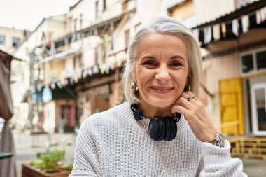 A happy mature woman wearing headphones smiles warmly while relaxing in a lively courtyard. clipart